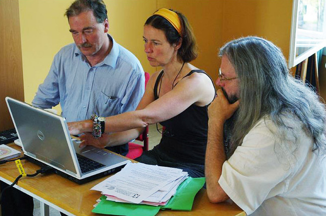 Three people sat at a table, discussing the content of a computer screen, at which the person in the middle is gesturing